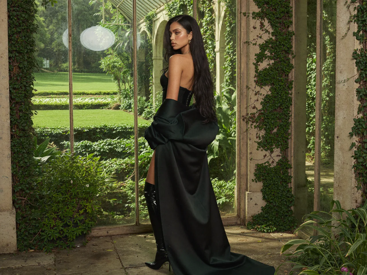 Seven Yuno in a black gown stands in a glasshouse doorway.